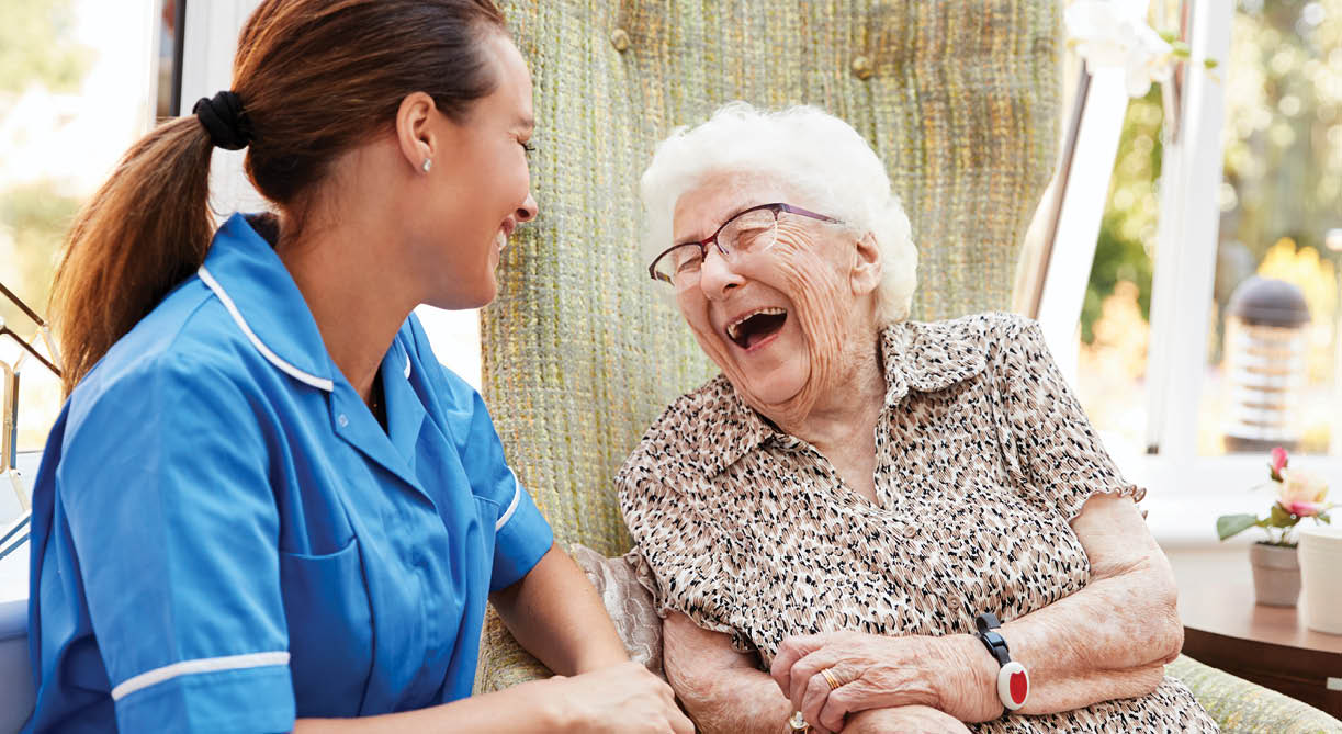 Senior Woman Sitting In Chair And Laughing With Nurse In Retirement Home