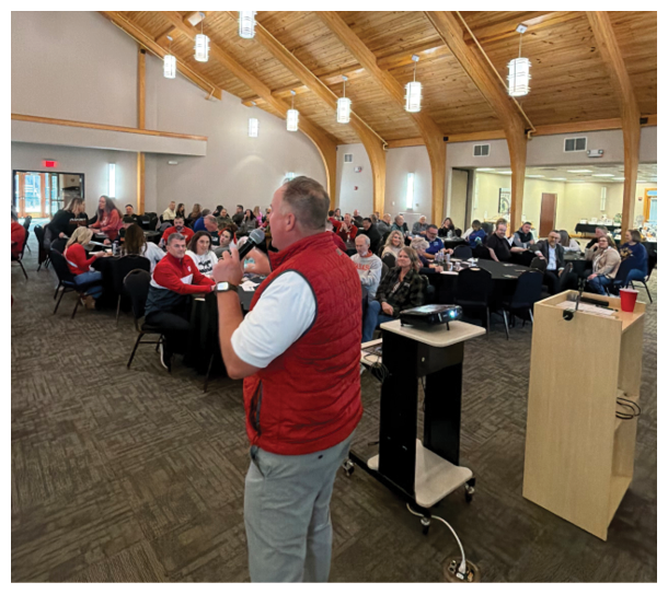 A man standing in a room with a red and white vest. Description generated by AI
