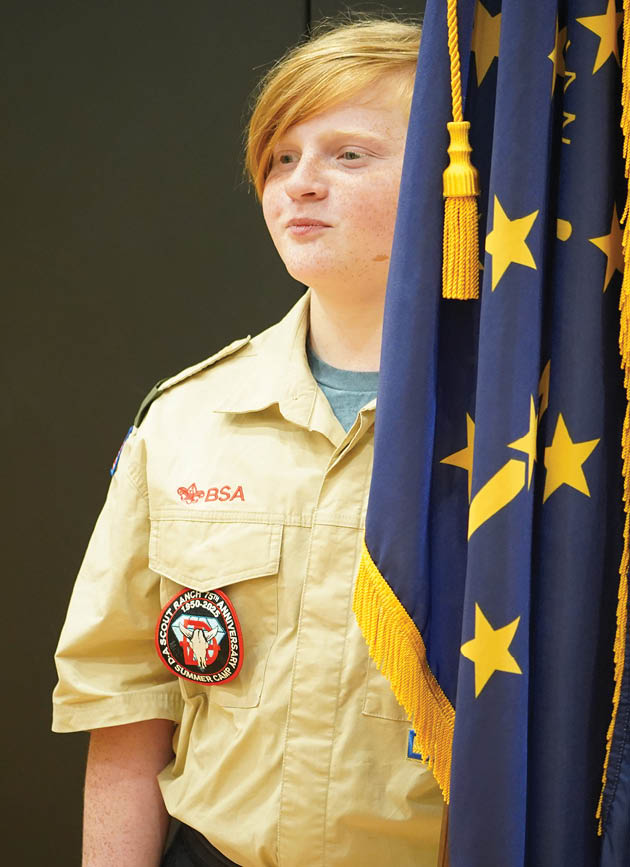 A young girl wearing a scout uniform stands in front of a flag. Description generated by AI