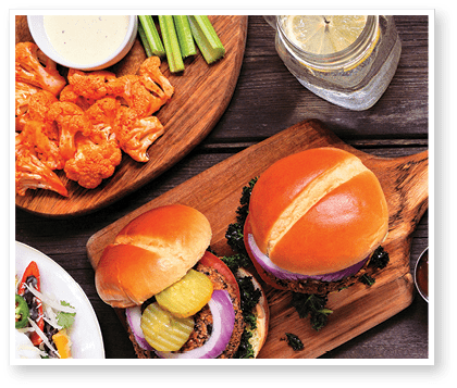 Healthy plant based fast food side border. Above view over a dark wood background. Table scene with cauliflower crust pizza, bean burgers and vegetarian sides. Copy space.