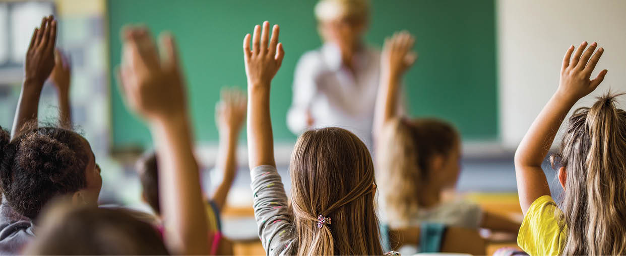 Rear view of large group of students raising their arms to answer the question on a class at elementary school.
