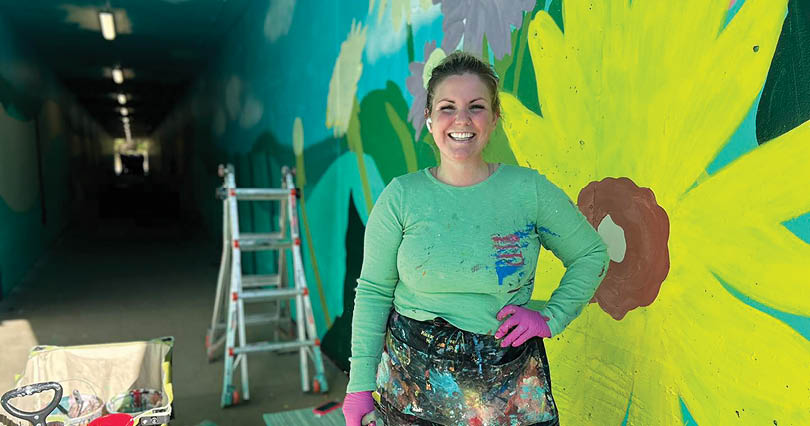 A woman is standing in front of a yellow flower painted on a wall. Description generated by AI