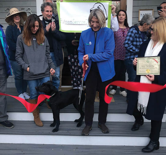 A group of people are gathered outside a house, with a woman in a blue jacket holding a pair of scissors. A black dog is also present, wearing a harness. The people are holding ribbons, and it appears that they are about to cut them. AI generated content