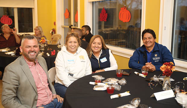 Four people are sitting around a table, posing for a picture. They are smiling and appear to be enjoying their time together. The table is set with various items, including wine glasses, cups, and a knife. There are also chairs placed around the table. AI generated content