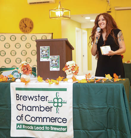 A woman is speaking at a podium in front of a table with a microphone. The table has a sign on it that says “Brewster Chamber of Commerce". AI generated content