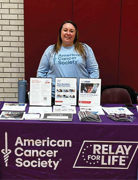 A woman stands behind a purple table with a sign that says “Relay for Life." AI generated content