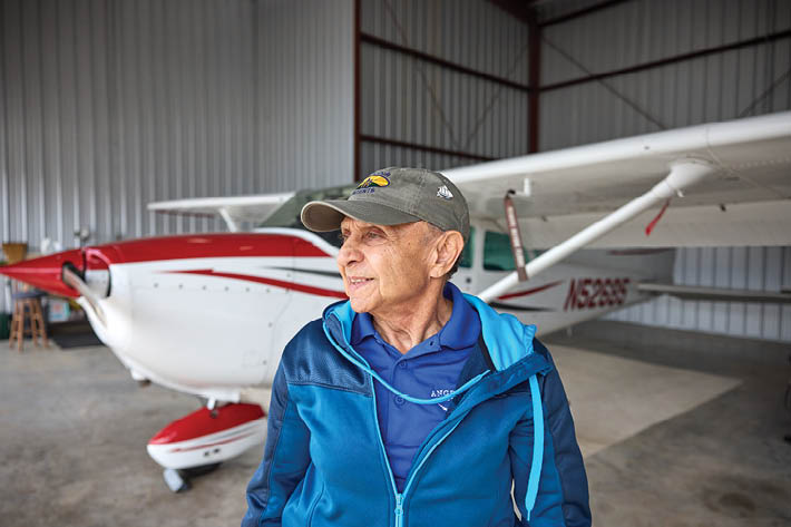 An older man wearing a blue jacket stands next to a white and red airplane. AI generated content