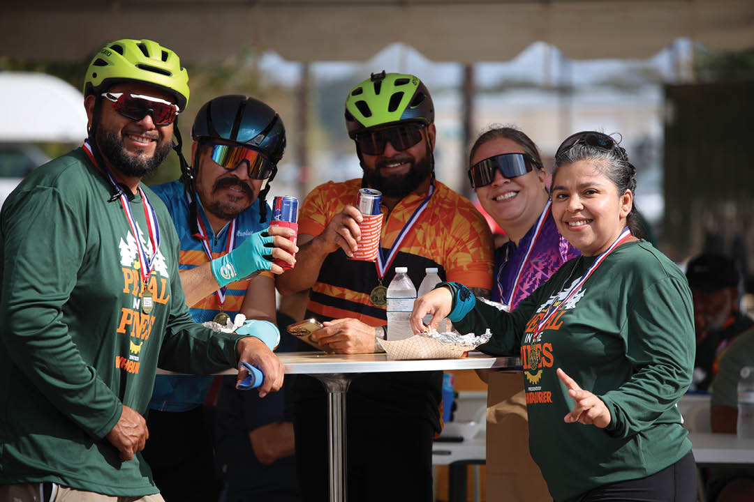 A group of people are sitting around a table, enjoying a meal together. They are wearing helmets, indicating that they might be participating in a cycling event. There are several bottles on the table, possibly containing water or other beverages. A cell phone can be seen on the table as well. AI generated content
