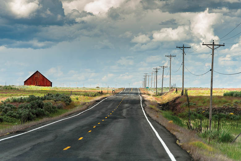 A lonesome highway in the Palouse area of eastern Washington State seems to lead to nowhere. A red barn can be seen in the distance. Power lines line the highway.