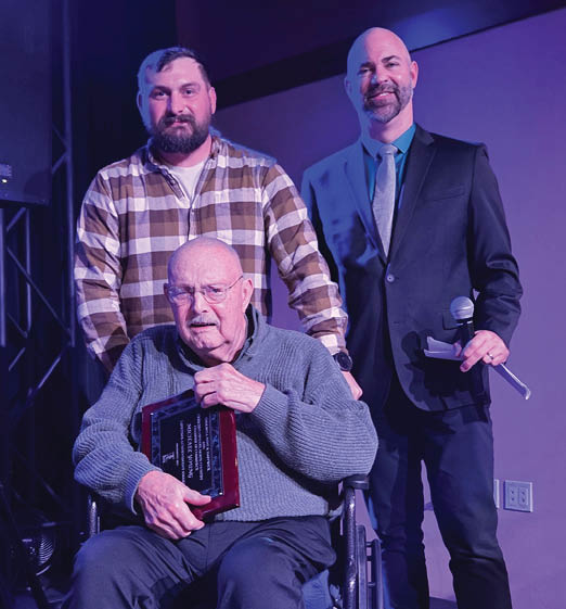 Three men standing on stage, one of them is in a wheelchair holding an award. AI generated content
