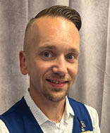 A man wearing a blue vest and a name tag sits in front of a curtain. AI generated content