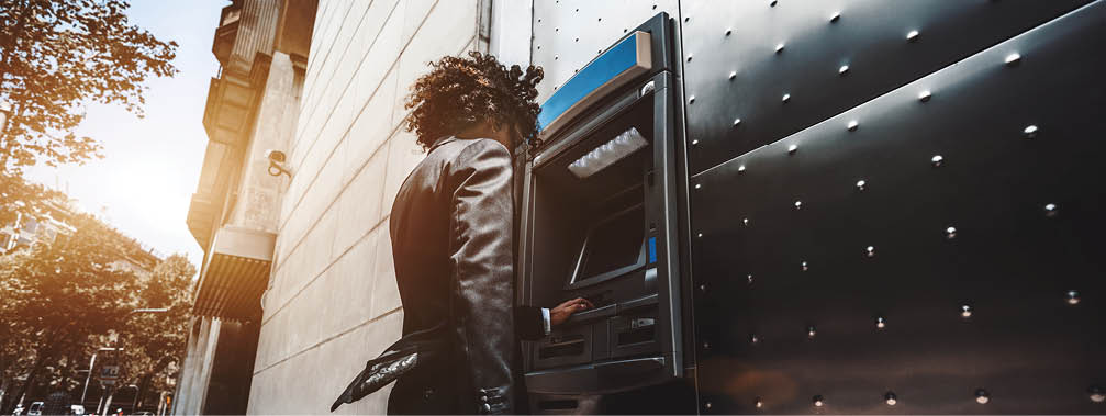 Wide-angle view of a curly businessman in a formal suit using an outdoor cash dispenser to increase his account; man entrepreneur is withdrawing money using a street ATM in urban settings
