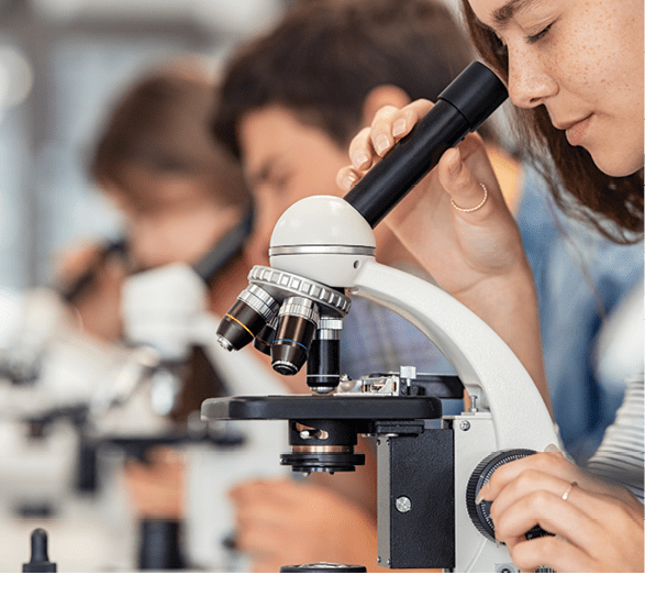 Close up of young woman seeing through microscope in science laboratory with other students. Focused college student using microscope in the chemistry lab during biology lesson. University students studying in high school science laboratory.