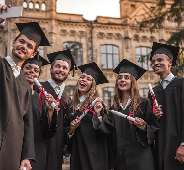 Successful graduates in academic dresses are holding diplomas, looking at camera and smiling while standing outdoors, guy is taking photo of them