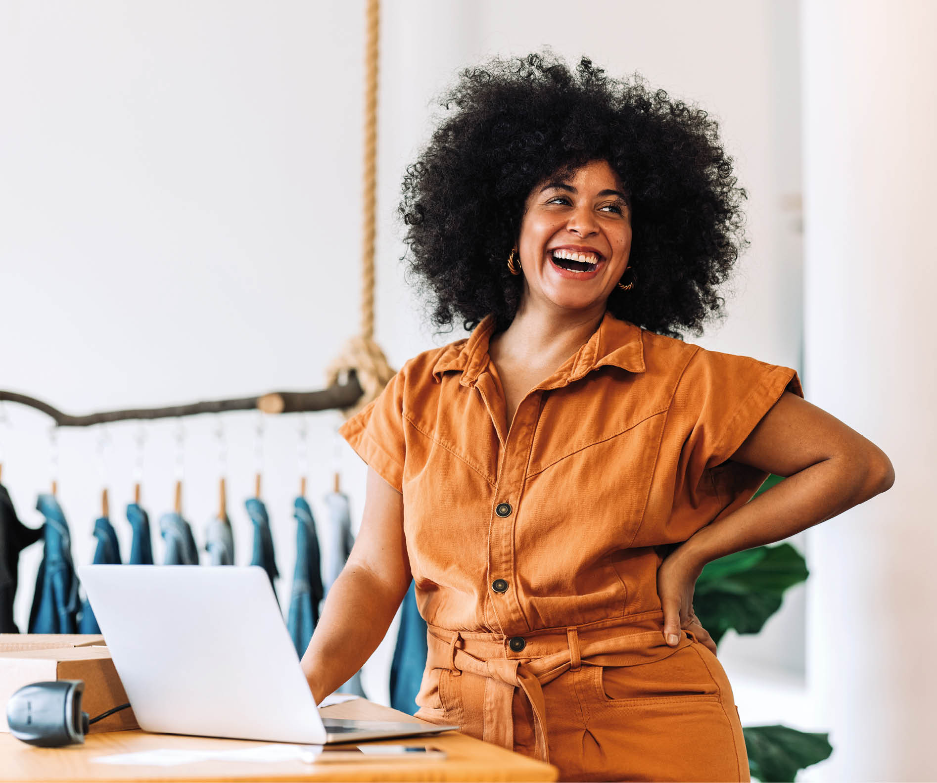 Ethnic small business owner smiling cheerfully while standing in her shop. Happy businesswoman managing her clothing orders on a laptop. Black female entrepreneur running an online clothing store.