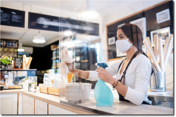 Portrait of young woman with face mask working indoors in cafe, disinfecting counter 