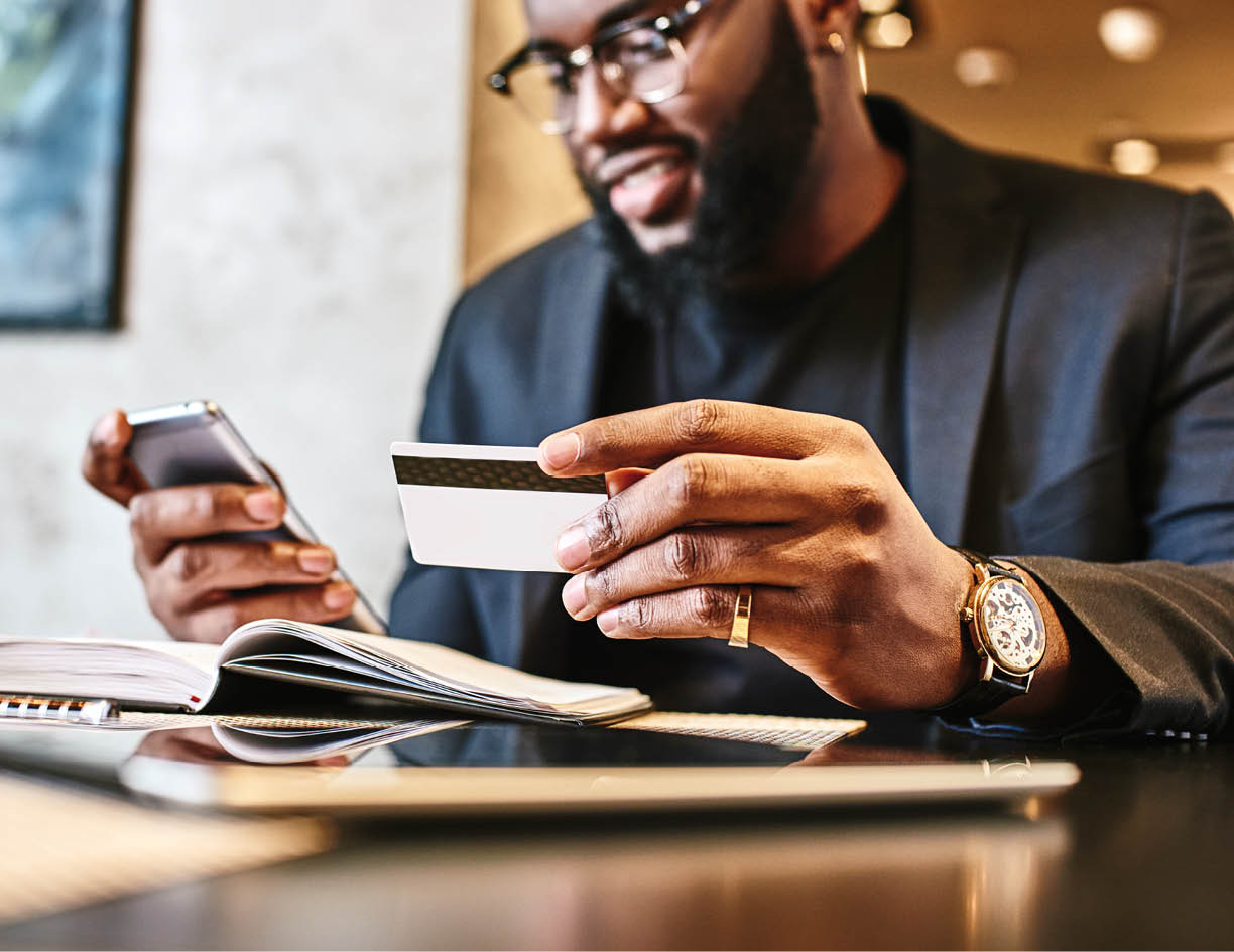 Portrait of African-American male holding cell phone in one hand and credit card in other, making transaction, using mobile banking app during lunch at cafe  His working papers and tablet pc are on the table  Online payment and shopping concept 