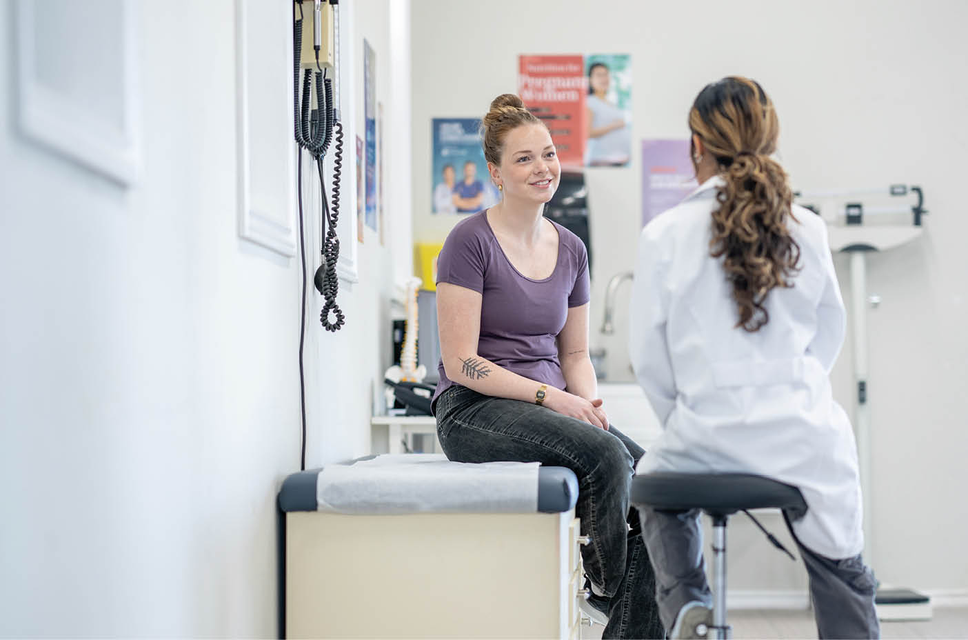 Two women are talking in a medical setting. One woman is sitting on a stool, and the other is standing. AI generated content