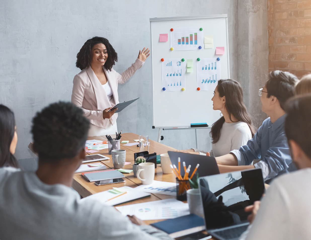 A woman is standing in front of a white board with a laptop and a cup on the table. She is pointing at the board while a group of people watch. AI generated content
