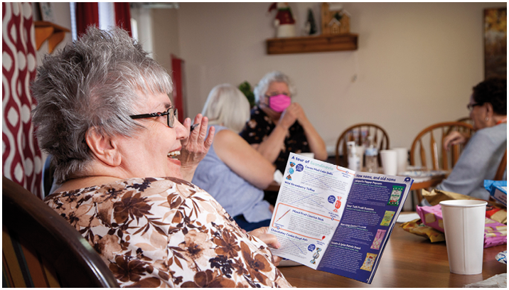 A group of older women are sitting around a table, smiling and enjoying each other's company. One woman is holding a magazine, possibly reading an article or looking at a map. The women are seated on chairs, with some of them holding cups. The scene appears to be a friendly gathering or a social event. AI generated content