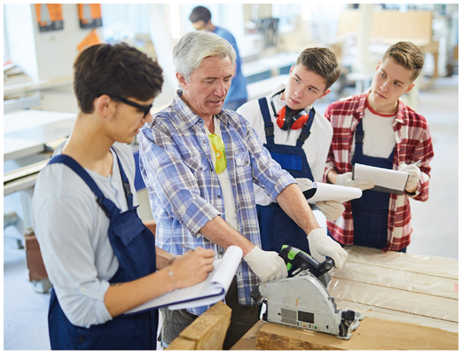 Serious experienced handsome mature carpenter in casual shirt standing at workbench and sawing wooden plank with circular saw at woodworking class, curious students listening to him