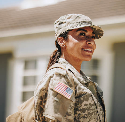 Happy female soldier looking away with a smile while standing outside her house with her bag. American servicewoman coming back home after serving her country in the military.