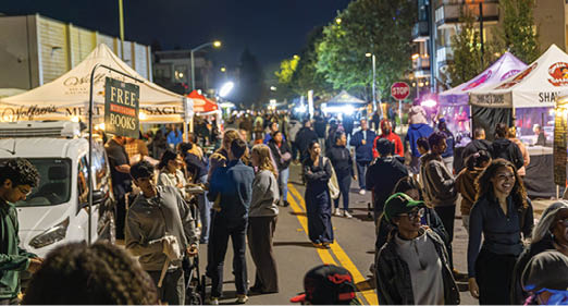 A large group of people are gathered on a street at night, with some standing in the street and others on the sidewalk. The crowd is diverse, with people of different ages and genders. There are also a few handbags and a backpack visible among the crowd. The street is lined with buildings, and a stop sign can be seen in the background. The overall atmosphere appears to be lively and social, with people enjoying their time together. AI generated content