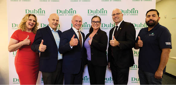 A group of six people pose for a photo in front of a white backdrop. They are all wearing suits and ties, with some of them giving thumbs up. The group consists of three men and three women, all standing close to each other. AI generated content