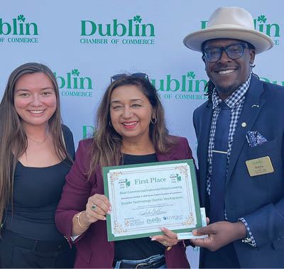 Three people are standing in front of a blue backdrop. A woman is holding a certificate, and a man is wearing a suit and tie. The man is smiling, and the woman is holding the certificate. AI generated content