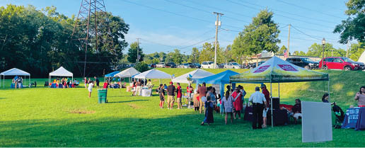 A group of people are gathered in a field, some sitting and some standing. There are several umbrellas set up, providing shade for the attendees. A few cars are parked in the background, and a truck can be seen further away. The scene appears to be an outdoor event or gathering. AI generated content
