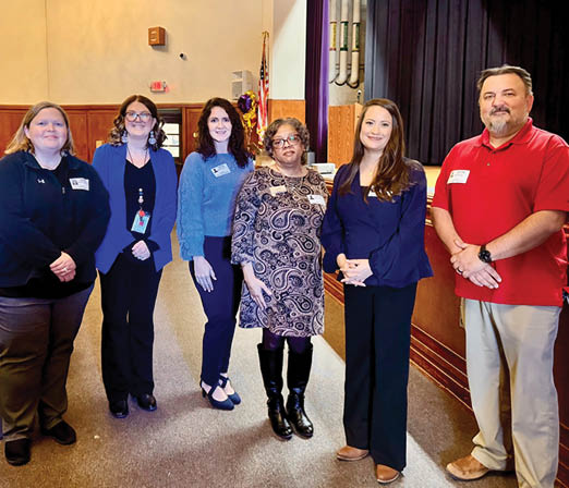 A group of six people, three men and three women, stand together in a hallway. They are all wearing name tags, indicating that they might be attending an event or gathering. The group appears to be posing for a picture, with some of them standing closer to the camera than others. AI generated content