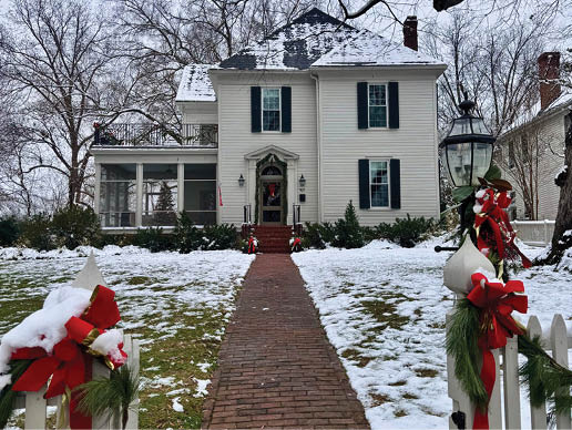 A large white house with a brick walkway leading to it. The house has a snowy roof and a wreath on the door. AI generated content
