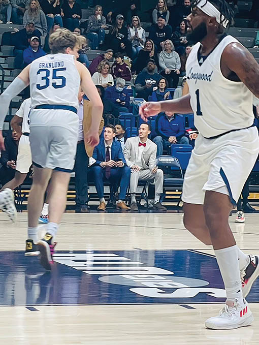 A basketball game is in progress with a man wearing a white jersey and white shorts. He is running down the court while another man in a blue jersey watches. The audience is seated in chairs surrounding the court, watching the game intently. AI generated content