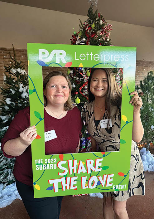 Two women pose for a picture in front of a green sign that says “Share the Love" and "The 2030 Subaru Share the Love Event". AI generated content