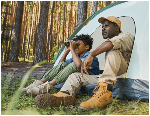 Cute African American boy looking through binoculars while sitting in tent next to his grandfather in casualwear during hike in the forest