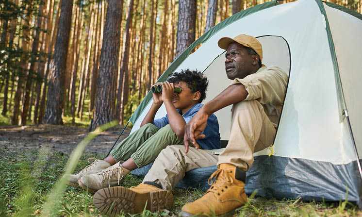Cute African American boy looking through binoculars while sitting in tent next to his grandfather in casualwear during hike in the forest