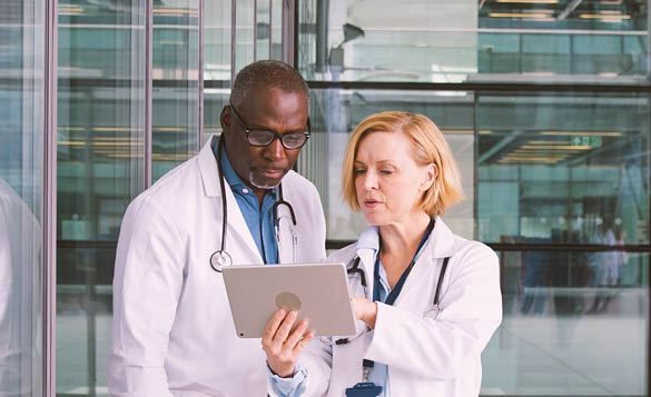 Male And Female Doctors Having Informal Meeting In Modern Hospital Looking At Digital Tablet