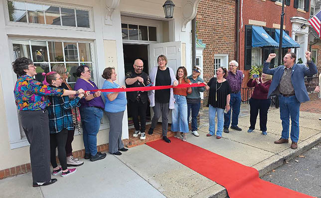 A group of people are gathered in front of a building, with a man holding a red ribbon. They are about to cut the ribbon, possibly for a grand opening or a special event. The people are standing on a red carpet, and some of them are wearing ties. The scene appears to be a celebration or a formal occasion. AI generated content