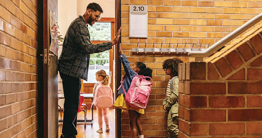 Elementary school teacher greeting his students at the door. Male teacher welcoming his class with a high five outside their classroom. Child mentor motivates his pupils in a primary school.