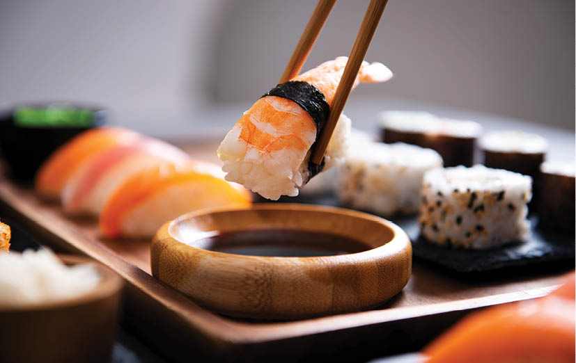 Closeup hand holding bamboo chopsticks with nigiri shrimp while soaking it in soy sauce. Detail of sushi set on wooden tray at restaurant while hand dip nigiri in soy sauce. Japanese cuisine concept.