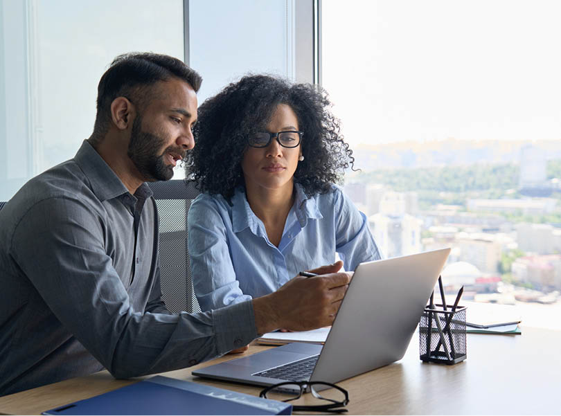 Indian male ceo executive manager mentor giving consultation on financial operations to female African American colleague intern using laptop sitting in modern office near panoramic window.