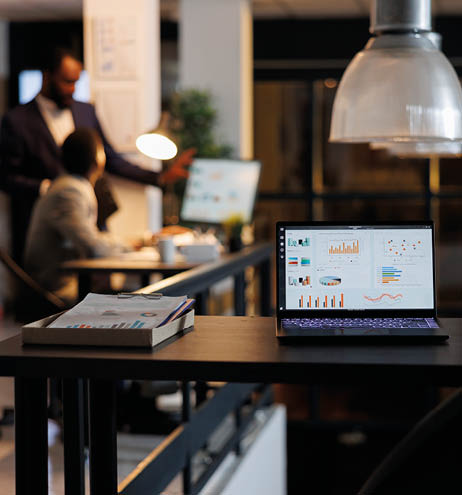 Laptop computer with financial graphs on screen, standing on table in startup office, coworkers discussing company strategy in background. Employees working late at night in startup office