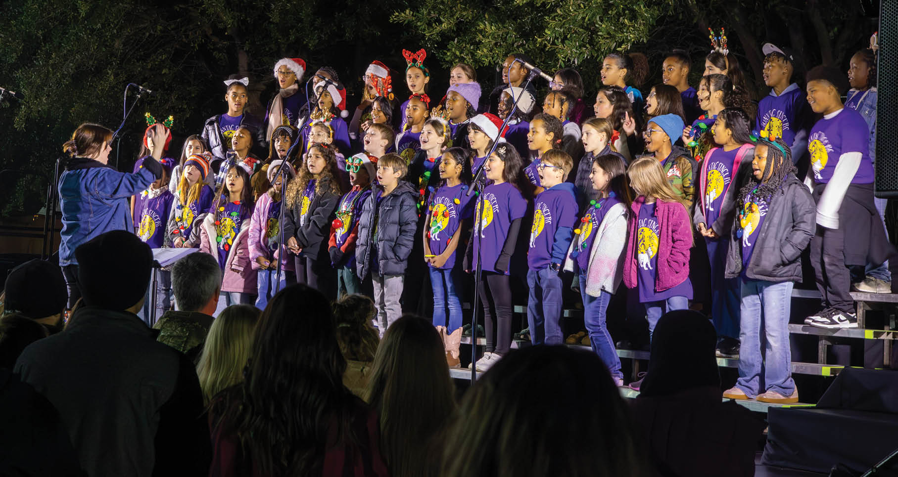 A group of children wearing Santa hats and purple shirts sing on stage. AI generated content