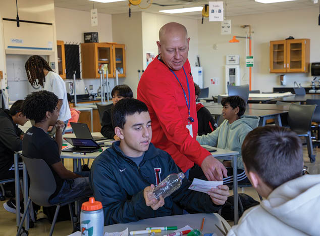 A man in a red shirt hands a video game controller to a young man in a classroom setting. AI generated content