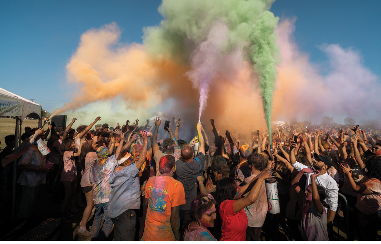 A large crowd of people are gathered together, with many of them holding up their arms. The sky is filled with colorful smoke, creating a vibrant and lively atmosphere. The people are enjoying the spectacle, with some of them even holding up their cell phones to capture the moment. AI generated content