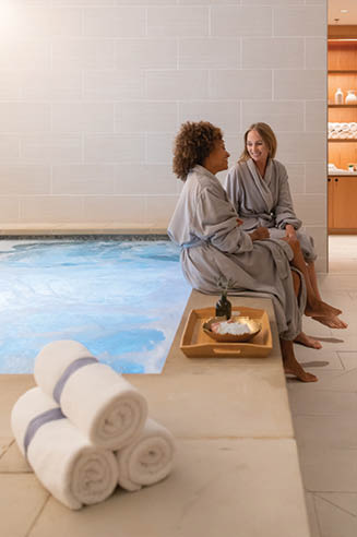 Two women are sitting in a spa, one on a bench and the other on a stool. They are both wearing robes. AI generated content