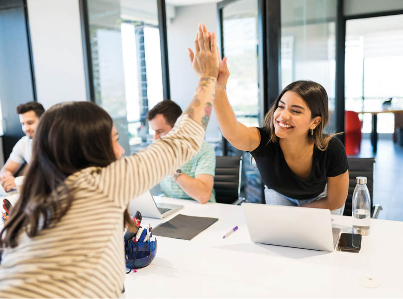 Successful female colleagues giving high-five at desk in creative workplace