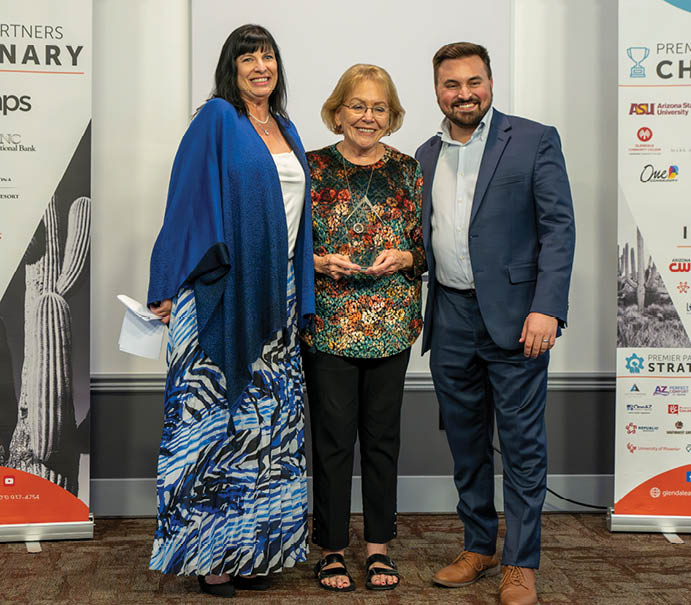 Three people are standing in front of a banner at a convention. A man and two women are posing for a picture. The man is wearing a suit and tie. AI generated content