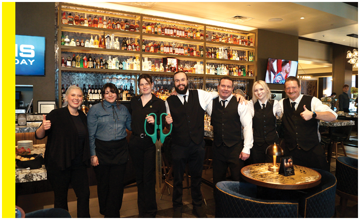 A group of six people, including a man and a woman, are standing in a restaurant. They are all wearing black and white clothing, and the woman is holding a pair of scissors. The group appears to be posing for a picture. AI generated content