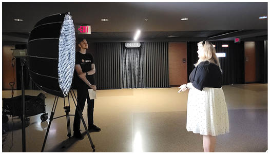 A man and a woman are standing in a room, with the woman wearing a black dress. The man is holding a camera, and they appear to be having a conversation. AI generated content